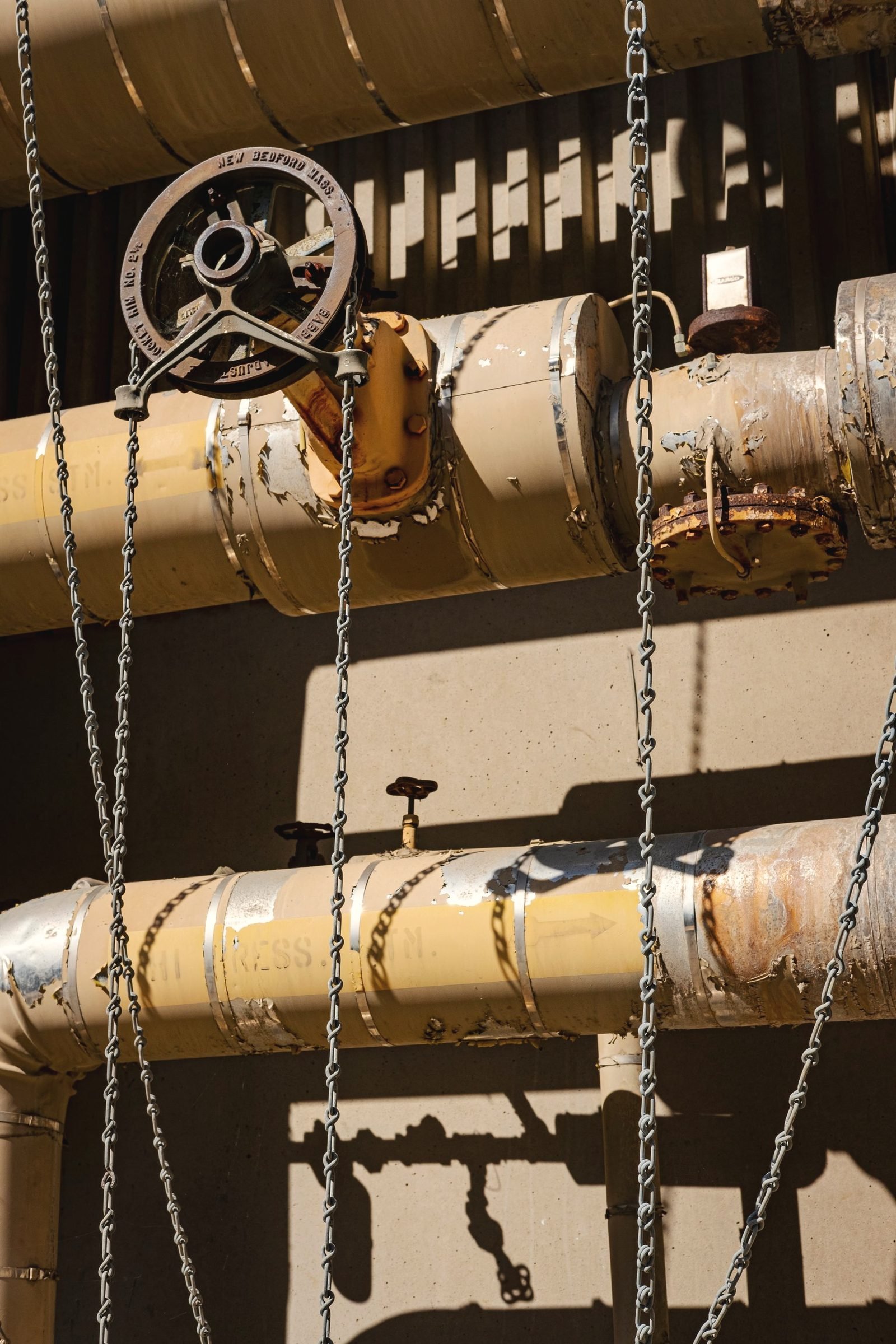 Wheel valve and hanging chains on old painted pipework in sunlight
